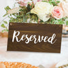 A dark wooden sign with the word Reserved written in white script, set against a backdrop of floral arrangements.