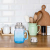 A clear blue glass mason jar shaker with a silver lid sits on a wooden table next to various kitchen tools and a large wooden