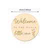 A round wooden sign with the text Welcome to the world little one engraved on it.