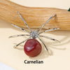 A handcrafted crystal spider figurine with silver legs and a reddish-orange carnelian bead resting on a wooden surface.
