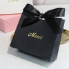 A black gift bag tied with a black ribbon featuring gold text that reads Merci, resting on a white surface.
