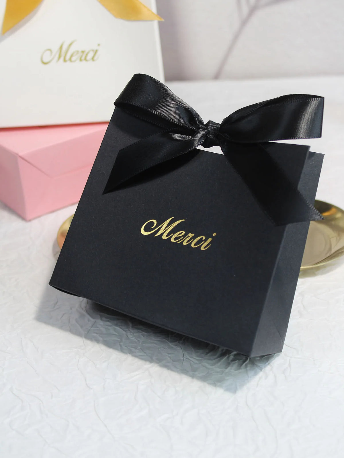 A black gift bag tied with a black ribbon featuring gold text that reads Merci, resting on a white surface.