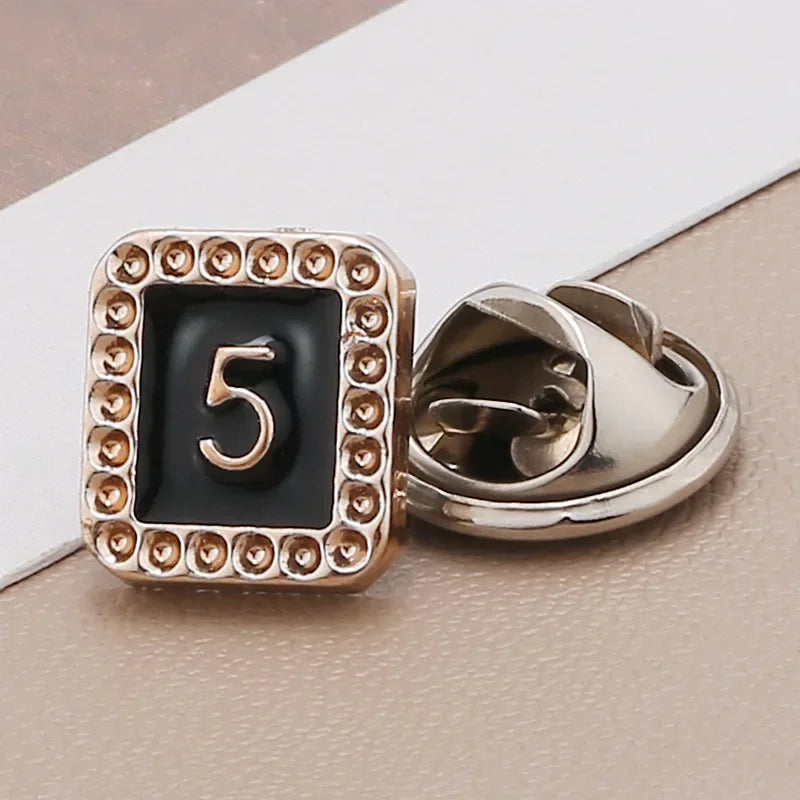 A close-up of a gold-toned square button with the number 5 engraved on it, set in a pearl and rhinestone border.