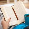 A hand is turning the page of an open book with a leather bookmark, resting on a blue fabric.