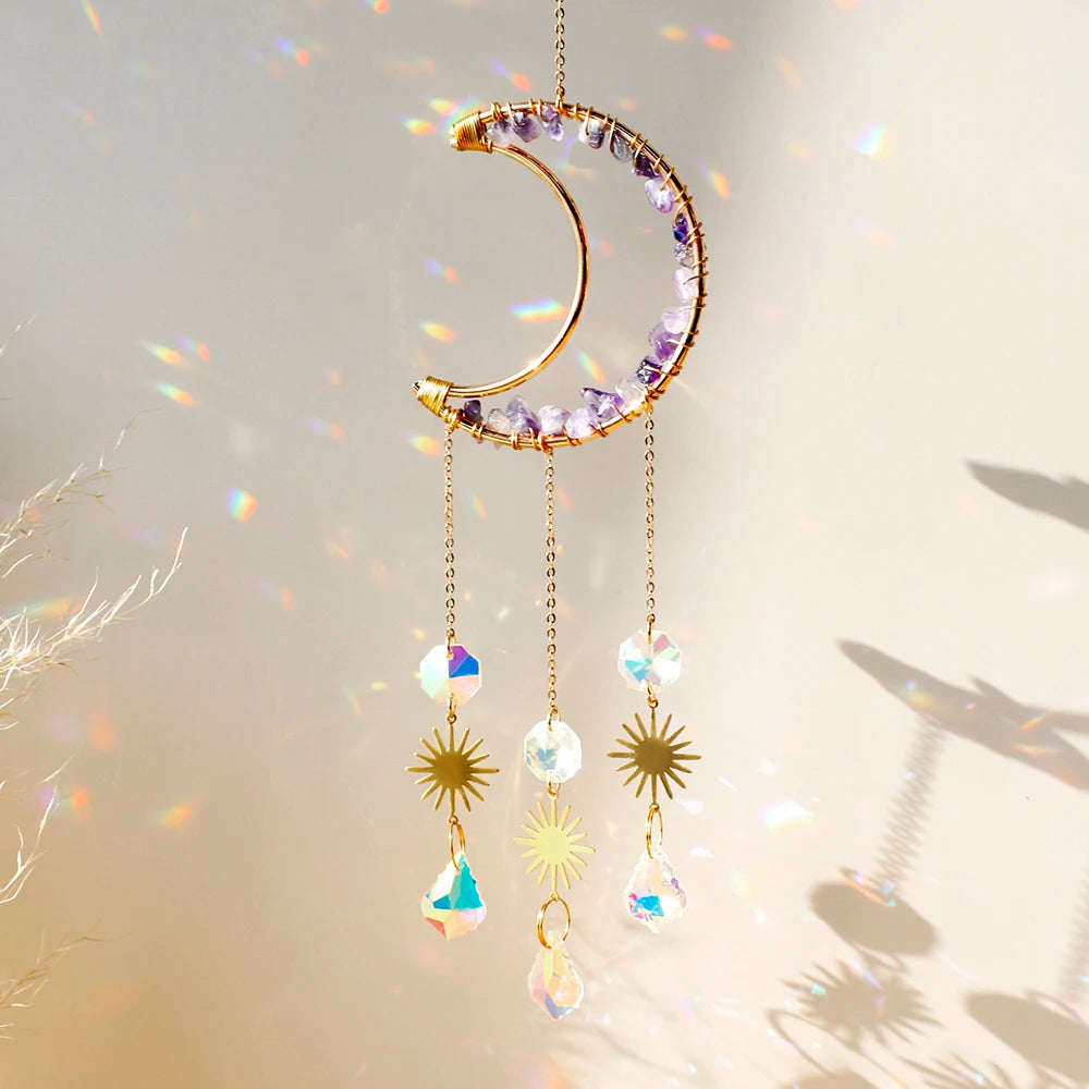 A golden crescent moon dreamcatcher with purple crystals and hanging prism charms against a soft light background.