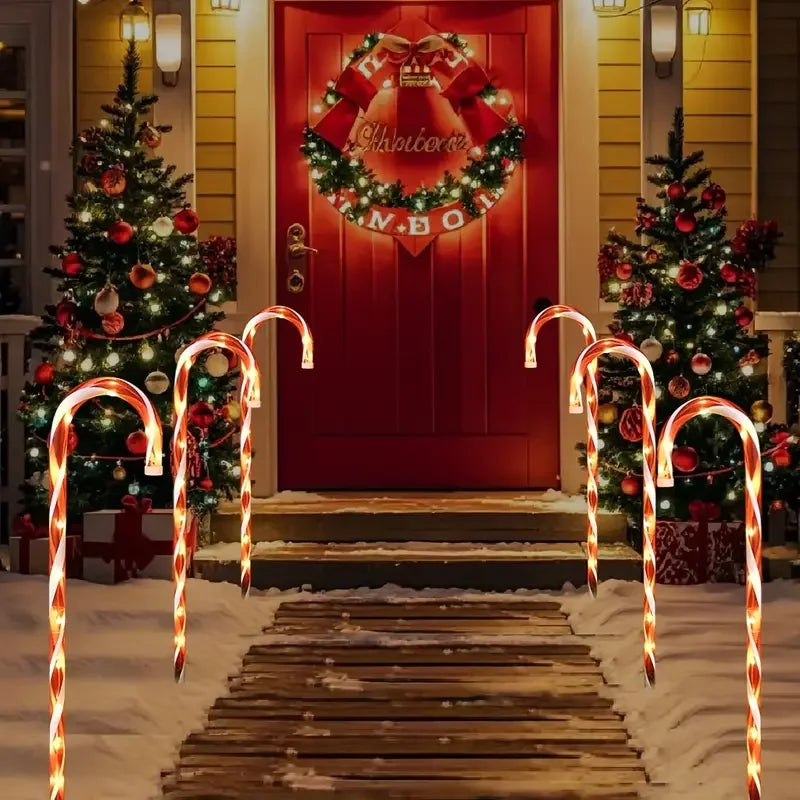 A pathway lined with glowing orange candy cane lights leads to a red door decorated with a Christmas wreath.