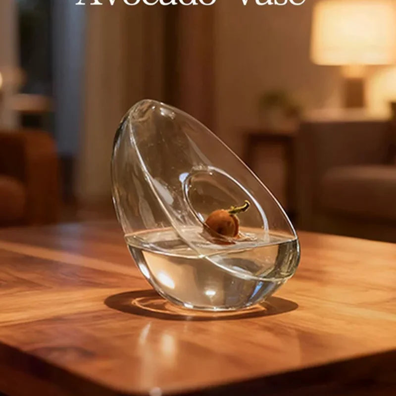 A clear glass vase shaped like an avocado seed is placed on a wooden table.