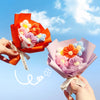 A hand holds an orange handmade crochet flower bouquet featuring pink, orange, and blue flowers.