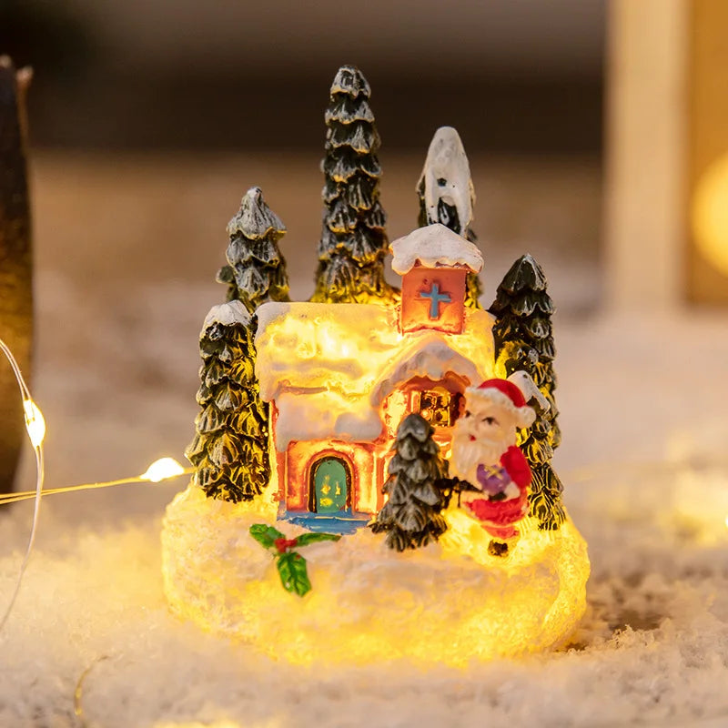 A glowing resin ornament depicting a Christmas gingerbread house surrounded by miniature evergreen trees and snow.