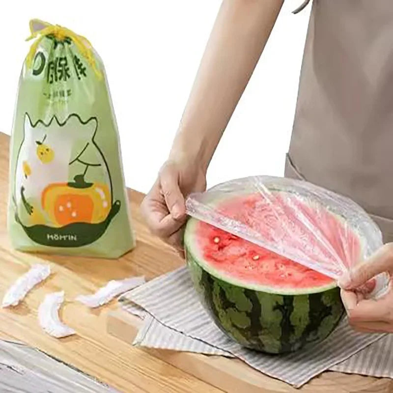 A hand is wrapping a slice of watermelon in a clear plastic bowl cover on a wooden surface.
