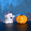 A white resin craft miniature ghost with purple accents next to an orange carved pumpkin.