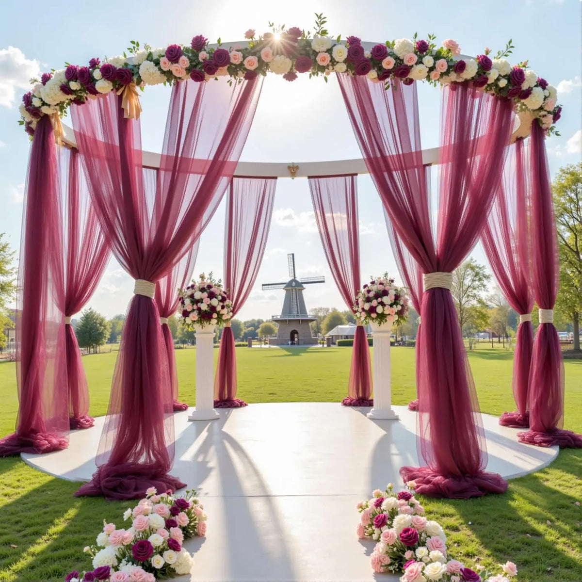 An elegant outdoor wedding backdrop featuring a pink floral arch and sheer tulle drapery.