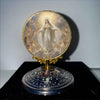 A golden framed religious ornament featuring a depiction of the Virgin Mary with a radiant halo and surrounding figures.