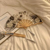 A leopard print folding hand fan with a black handle rests on a light-colored sheet, next to a wooden fan and a book.