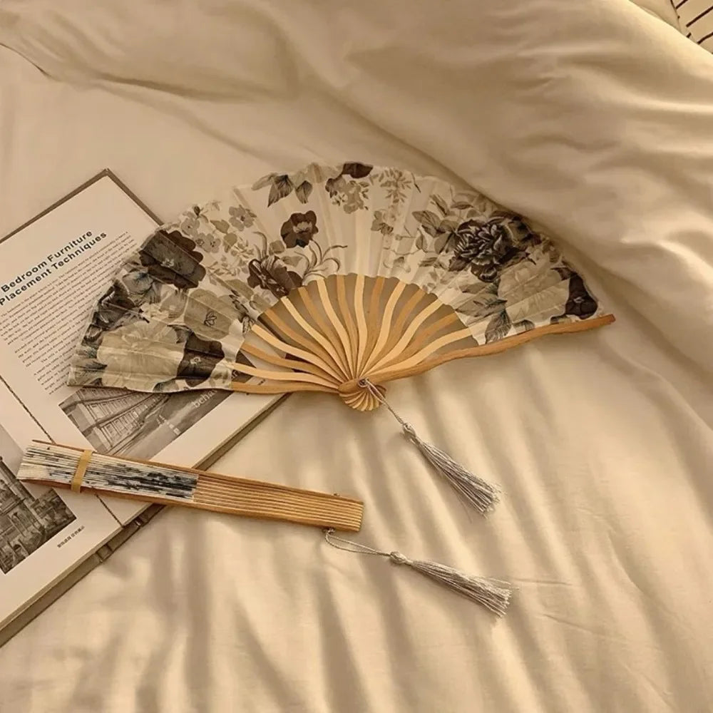 A leopard print folding hand fan with a black handle rests on a light-colored sheet, next to a wooden fan and a book.