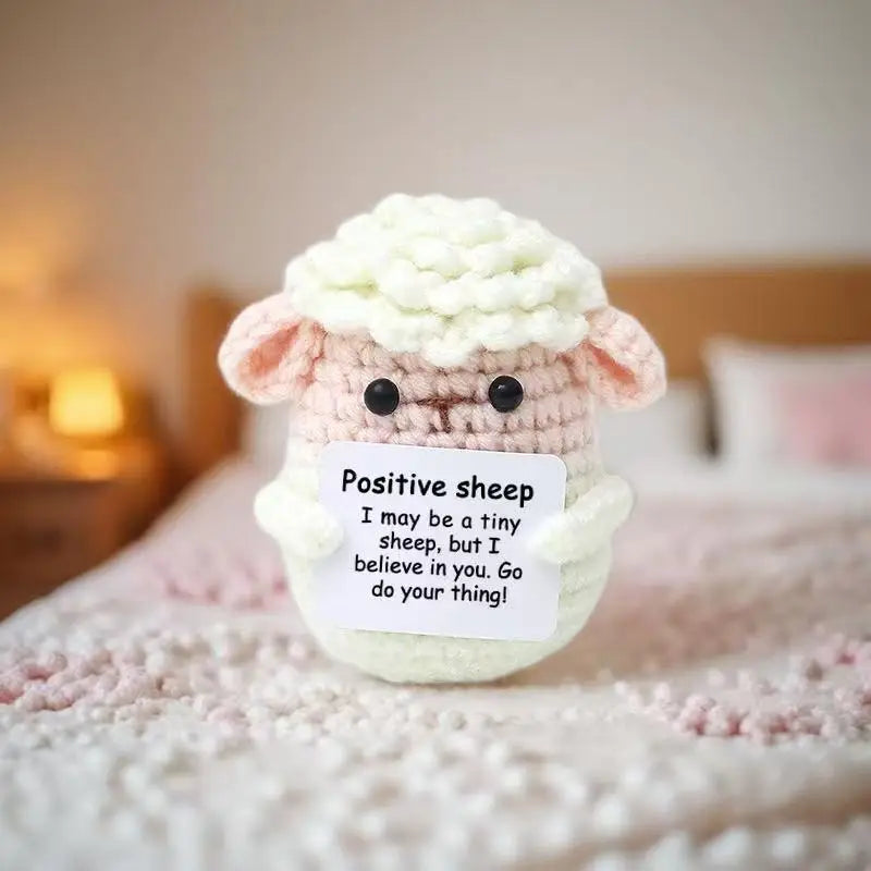Handmade crochet sheep baseball bat doll with a positive message card sitting on pink fabric.