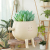 A light beige resin succulent planter shaped like a smiling creature is hanging from a wooden swing base.