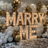 A display of marquee light-up letters spelling 'MARRY ME' decorated with gold and white balloons.