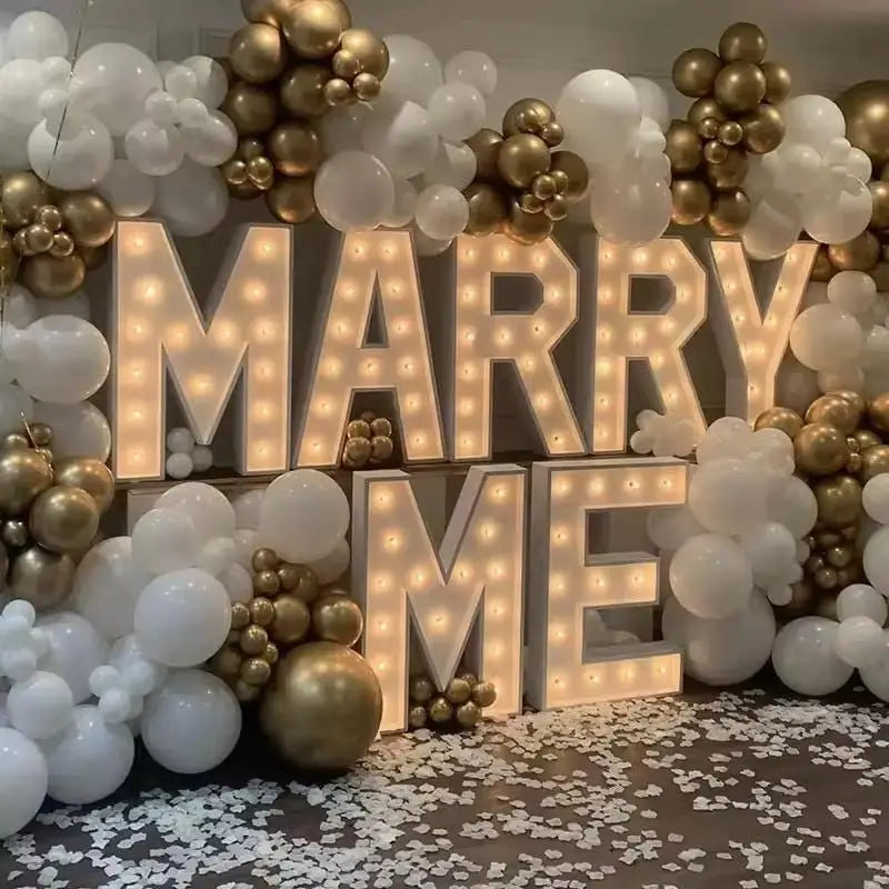 A display of marquee light-up letters spelling 'MARRY ME' decorated with gold and white balloons.