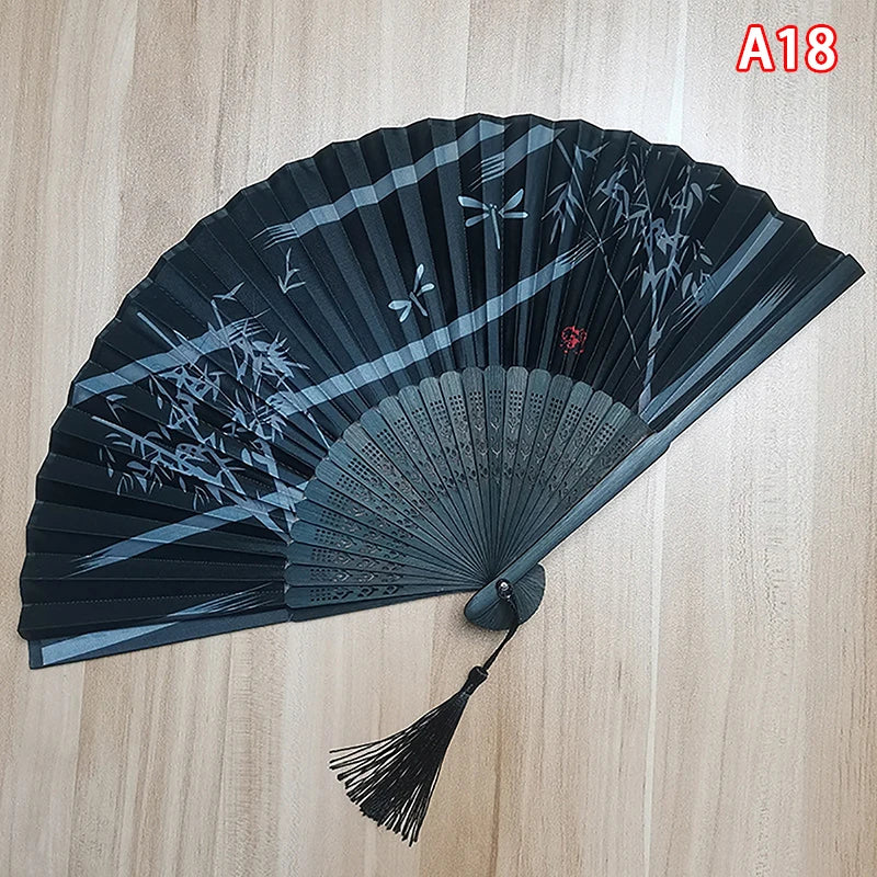 A dark gray folding hand fan with intricate black floral designs and a tassel.