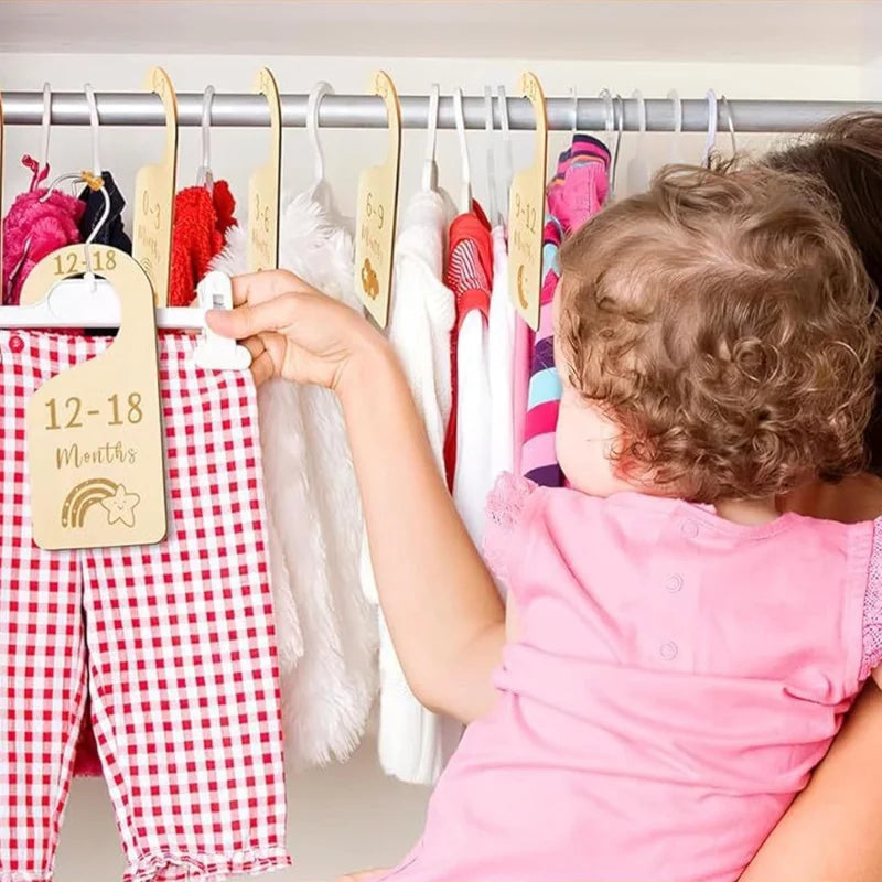 A young child is looking at a rack of baby clothes hanging on hangers with rainbow cloud-shaped dividers.
