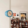 A blue modern resin wall clock with a white face and orange hands is mounted on a light gray wall above wooden shelving.