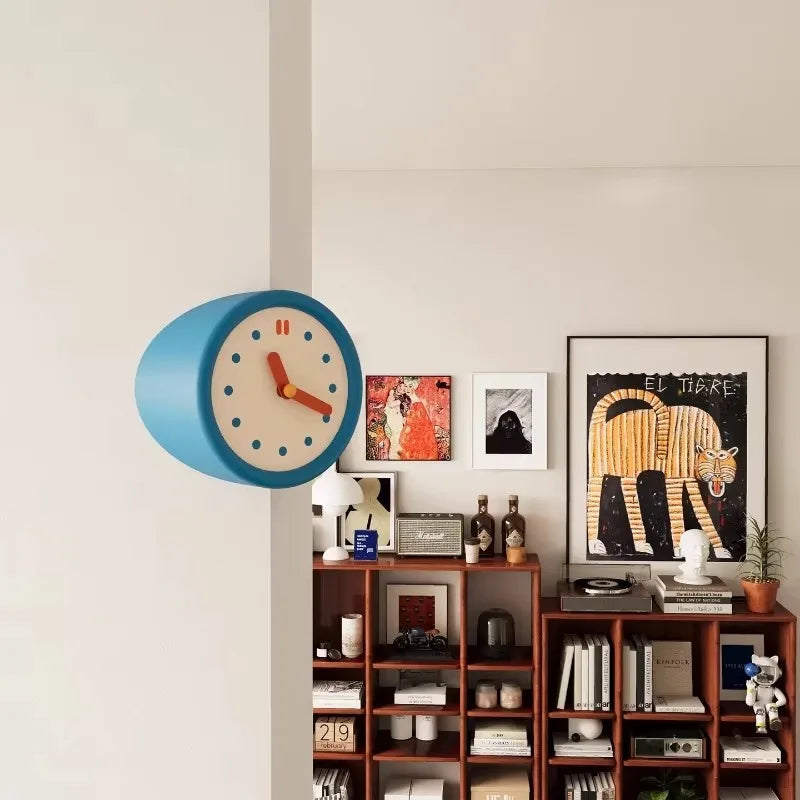 A blue modern resin wall clock with a white face and orange hands is mounted on a light gray wall above wooden shelving.