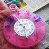 A pink and purple marbled vintage vinyl record wall clock with a white face is displayed on a surface next to a newspaper.