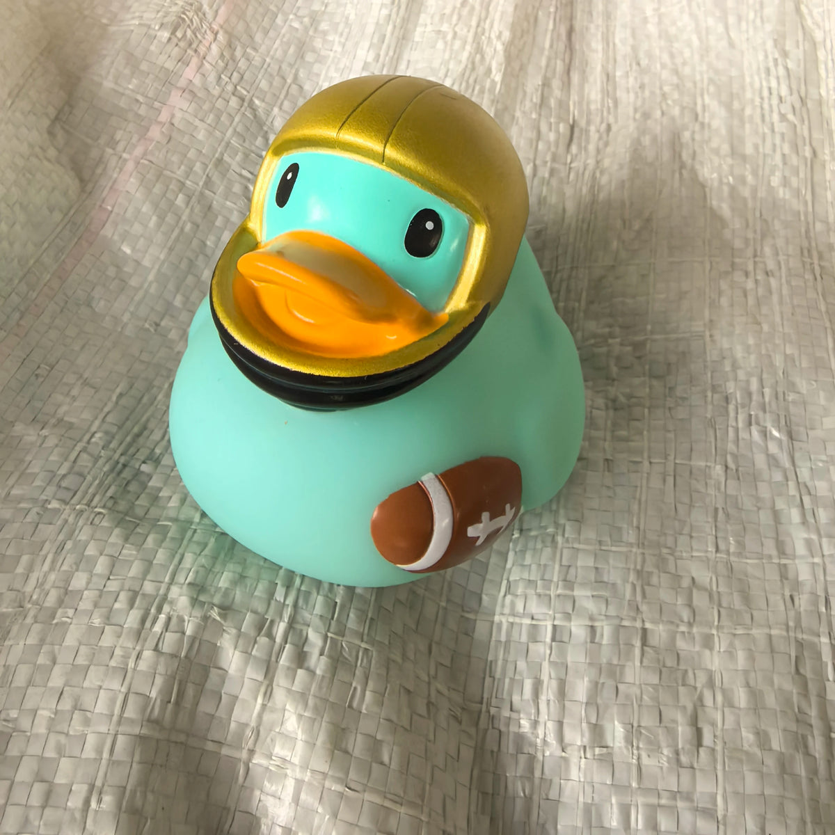 A bright turquoise rubber duck bath toy with a gold-colored head and an orange beak resting on a light gray textured surface.