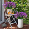 A white folding stand holds a large purple flower arrangement and several pumpkins in front of a green garden backdrop.