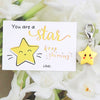 A card with the text You are a star keep shining and a yellow star keychain hanging from a silver ring amidst white flowers.