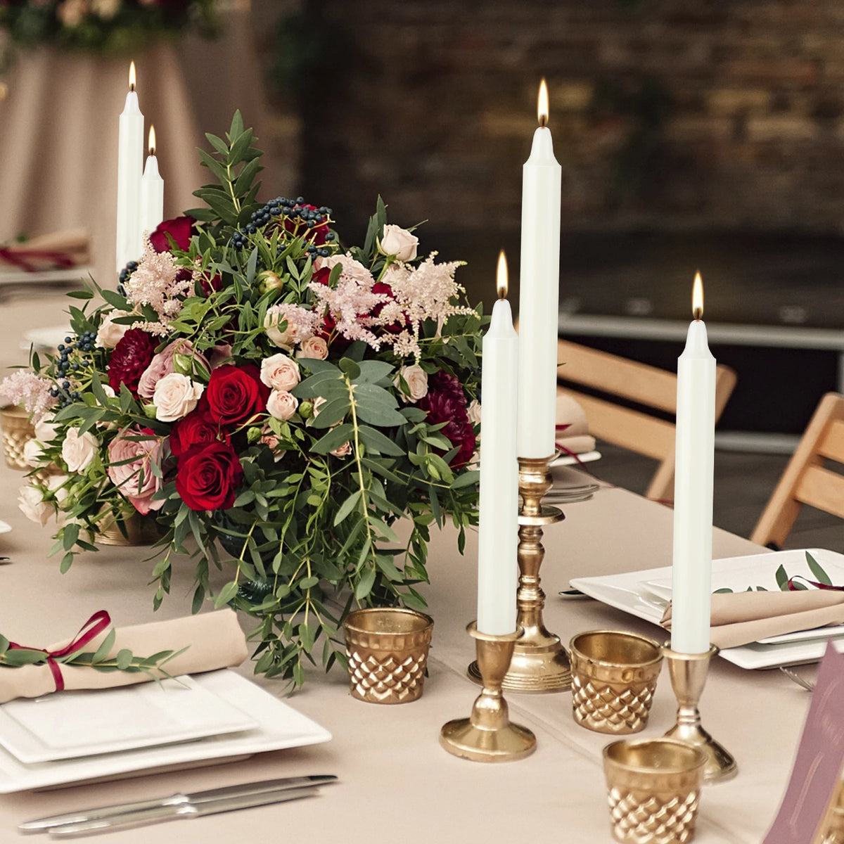 A floral arrangement featuring deep red and pink roses and greenery, set on a table with white candles.