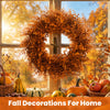 An artificial autumn wreath made of dried orange leaves hangs in front of a window with fall harvest decorations.