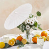A white decorative paper parasol umbrella is shown next to a bouquet of white flowers and citrus fruits on a white table.