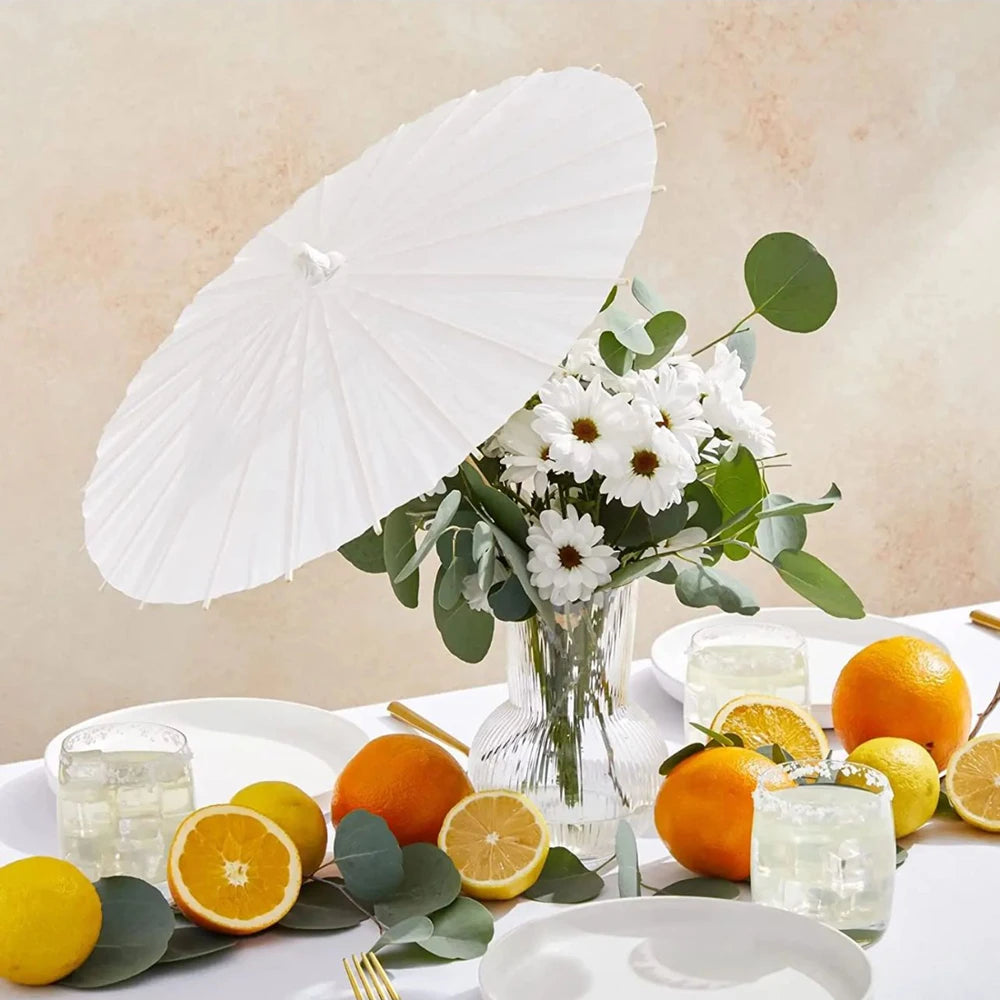A white decorative paper parasol umbrella is shown next to a bouquet of white flowers and citrus fruits on a white table.