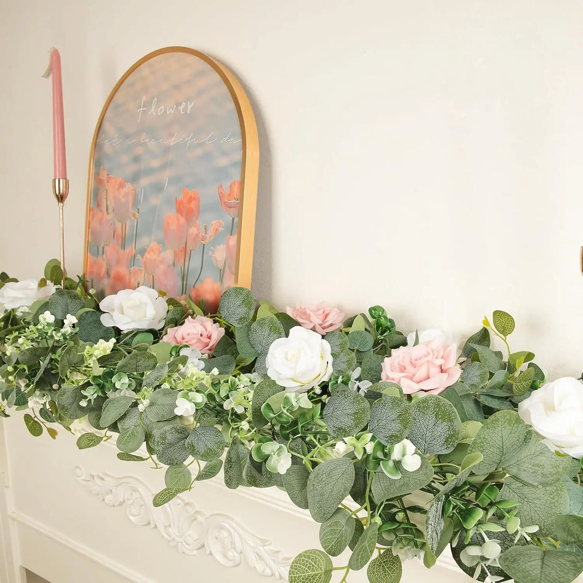 Guirlande artificielle d'eucalyptus avec des fleurs blanches et roses, exposée sur un mur avec une bougie rose.