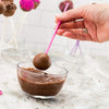 A hand holds a pink lollipop stick dipped in melted chocolate, hovering over a glass bowl of chocolate mixture.
