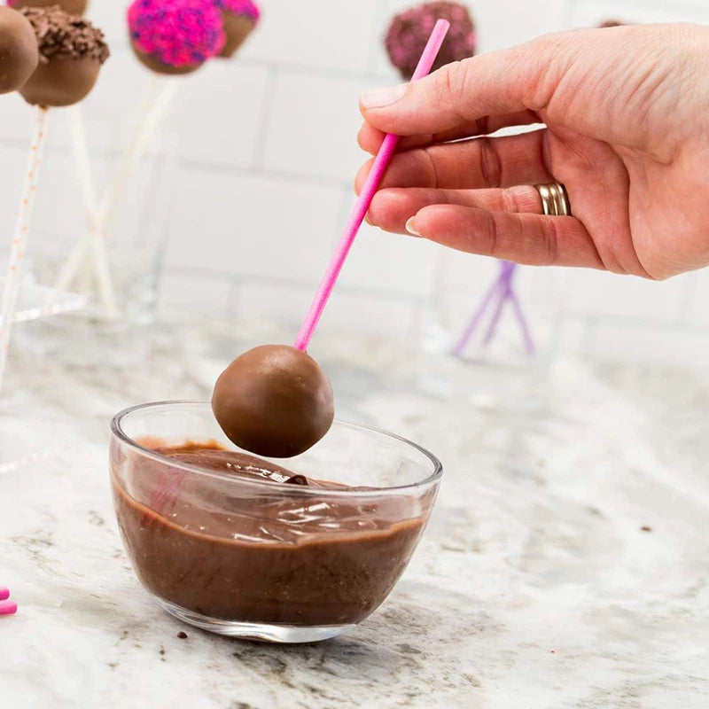 A hand holds a pink lollipop stick dipped in melted chocolate, hovering over a glass bowl of chocolate mixture.