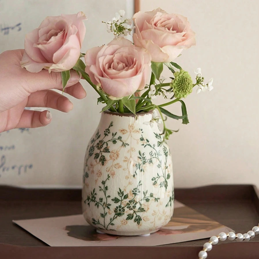 A hand holds a pink ceramic vase filled with light pink roses and green foliage.