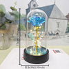 A glass dome featuring an eternal blue rose with a golden stem and LED light, displayed on a white surface.