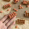 A hand holds a wooden egg carton filled with small, round, orange-colored eggs on a beige fabric surface.