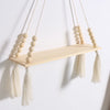 A rustic wooden wall shelf with macrame tassels hangs against a white wall.