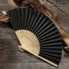 A black and light wood hand folding fan is displayed on a dark wooden surface.