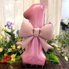 A large pink balloon shaped like the number one with a matte bow, displayed among greenery.