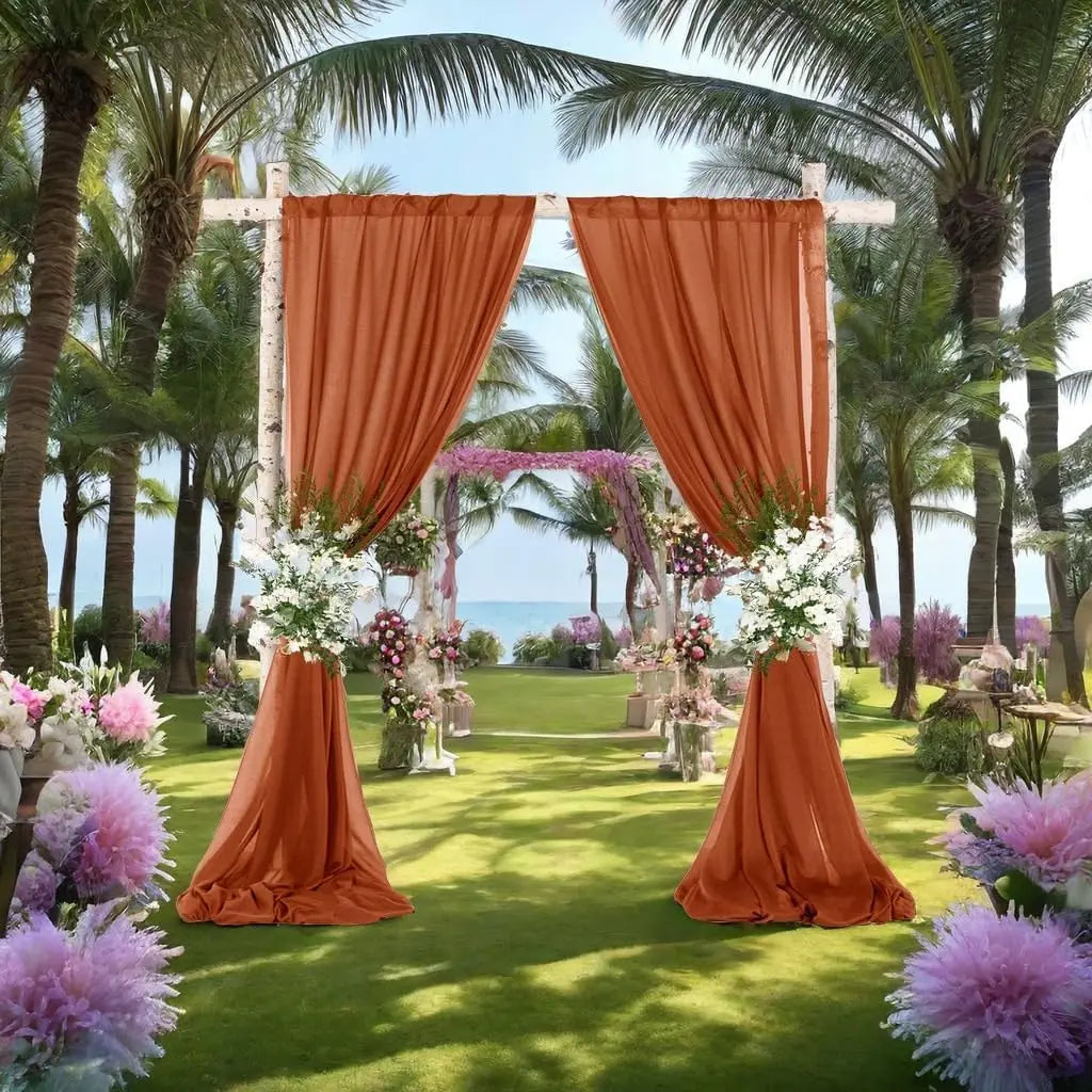 An outdoor wedding backdrop featuring orange chiffon curtains framing a lush garden and palm trees.