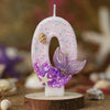 A white birthday candle shaped like the number 0 decorated with purple and iridescent glitter and a silver starfish flower.