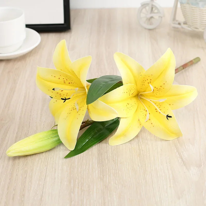 A realistic silk lily flower stem with three yellow blooms and green leaves on a wooden surface.