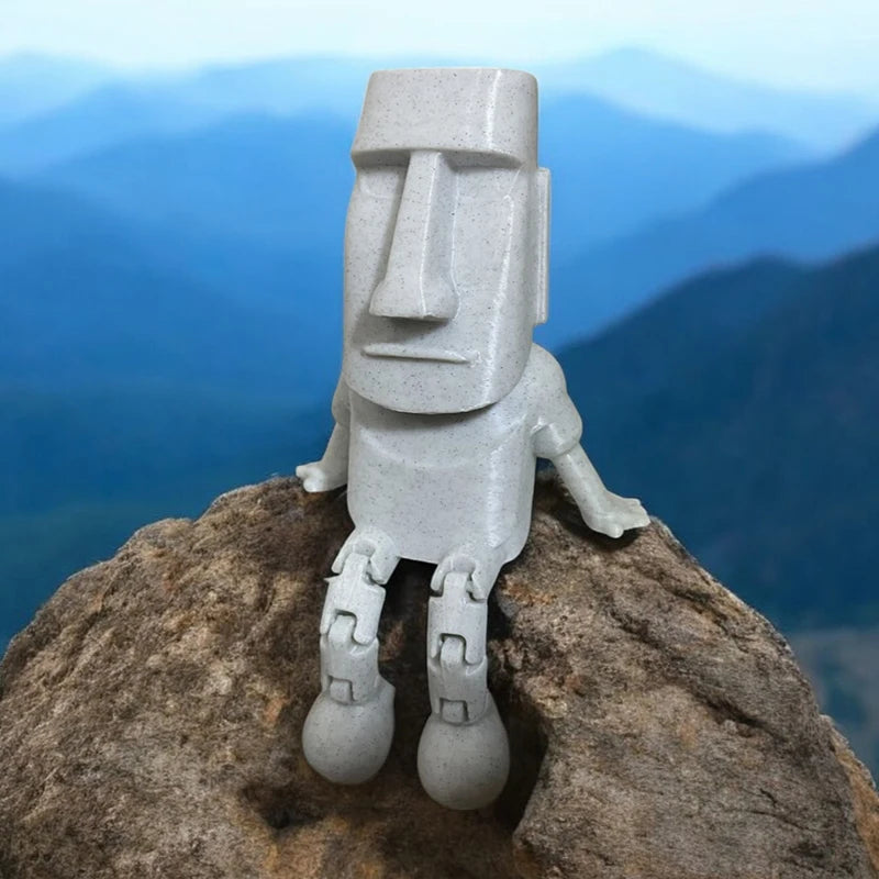 A gray 3D printed Moai statue figurine with movable joints standing on a large rock against a blue mountain background.