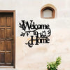 A black metal welcome sign with elegant script reading Welcome to Our Home mounted on a white stucco wall next to a wooden do