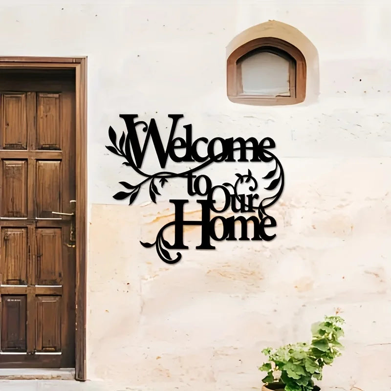 A black metal welcome sign with elegant script reading Welcome to Our Home mounted on a white stucco wall next to a wooden do